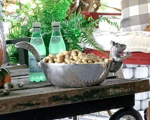 A decorative nut bowl viewed from the side, featuring a playful squirrel handle, filled with peanuts on a rustic table.