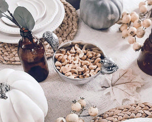 A nut bowl filled with cashews, viewed from above, sits amid autumn-themed decor on a woven placemat.
