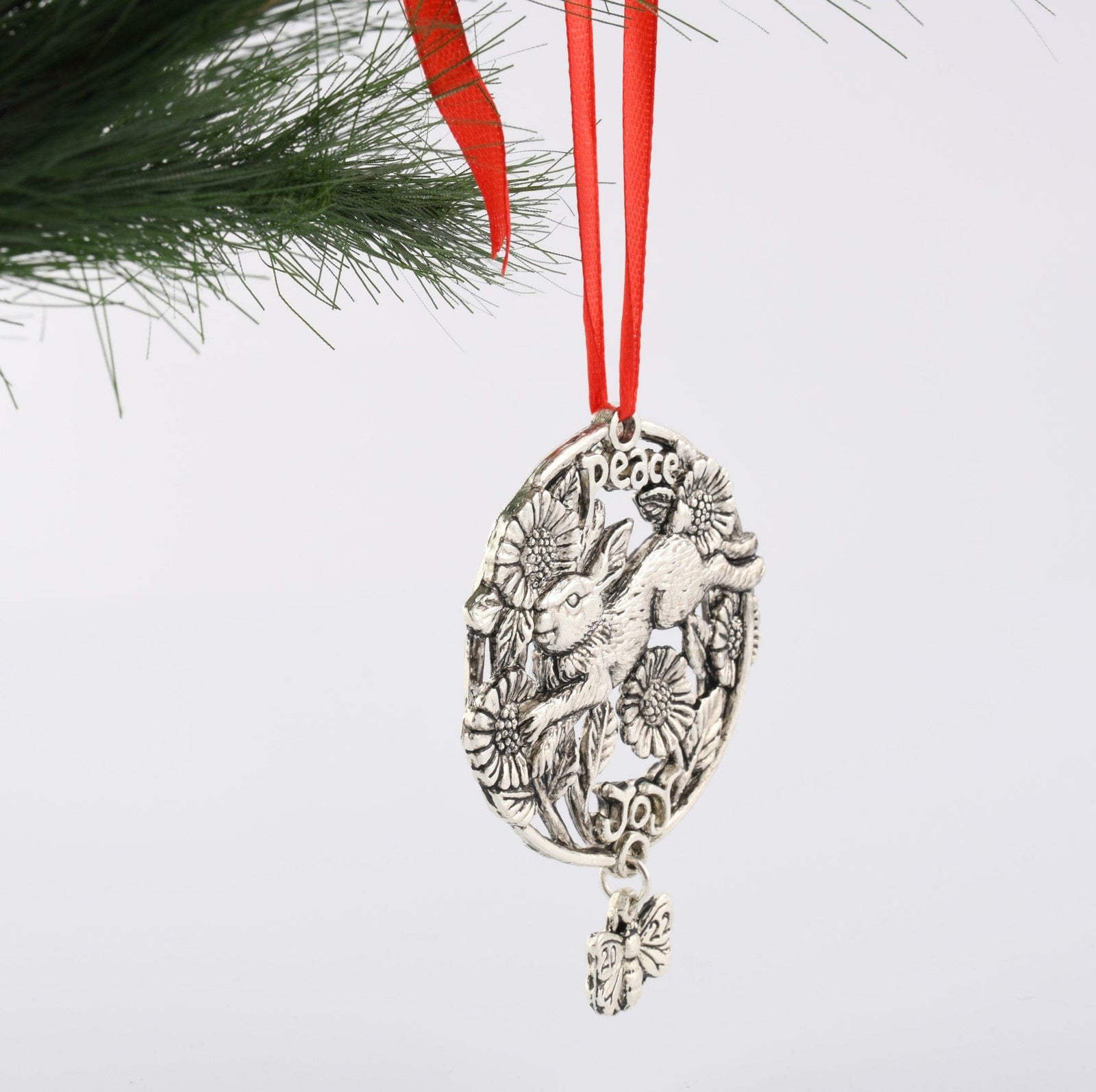 Hanging ornament with rabbit and floral details, accented by a red ribbon, seen from the side on a pine branch.