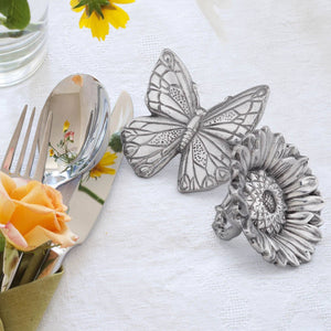 Butterfly and sunflower napkin rings on a white tablecloth, viewed from above, beside elegant silverware and fresh flowers.