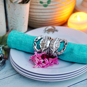 A coastal-themed napkin ring with crab design holding a teal napkin, styled elegantly on stacked plates and seen from a side angle.