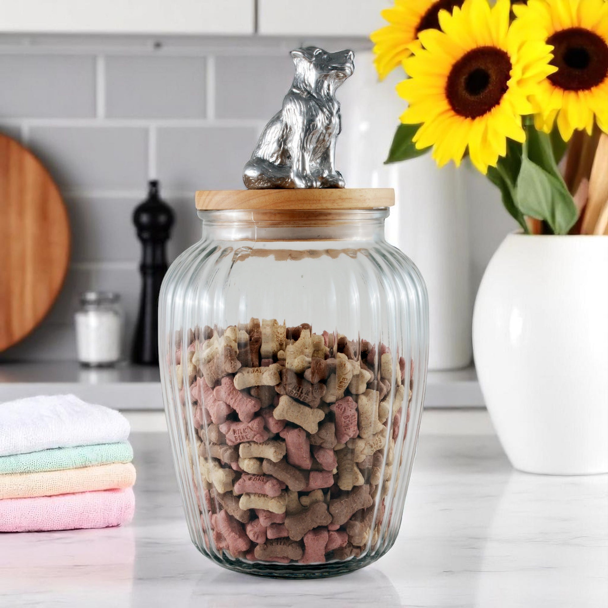 A glass canister filled with dog treats, viewed from a slightly elevated angle, features a wooden lid topped with a decorative silver dog, ideal for luxury home accessory and kitchen decor.