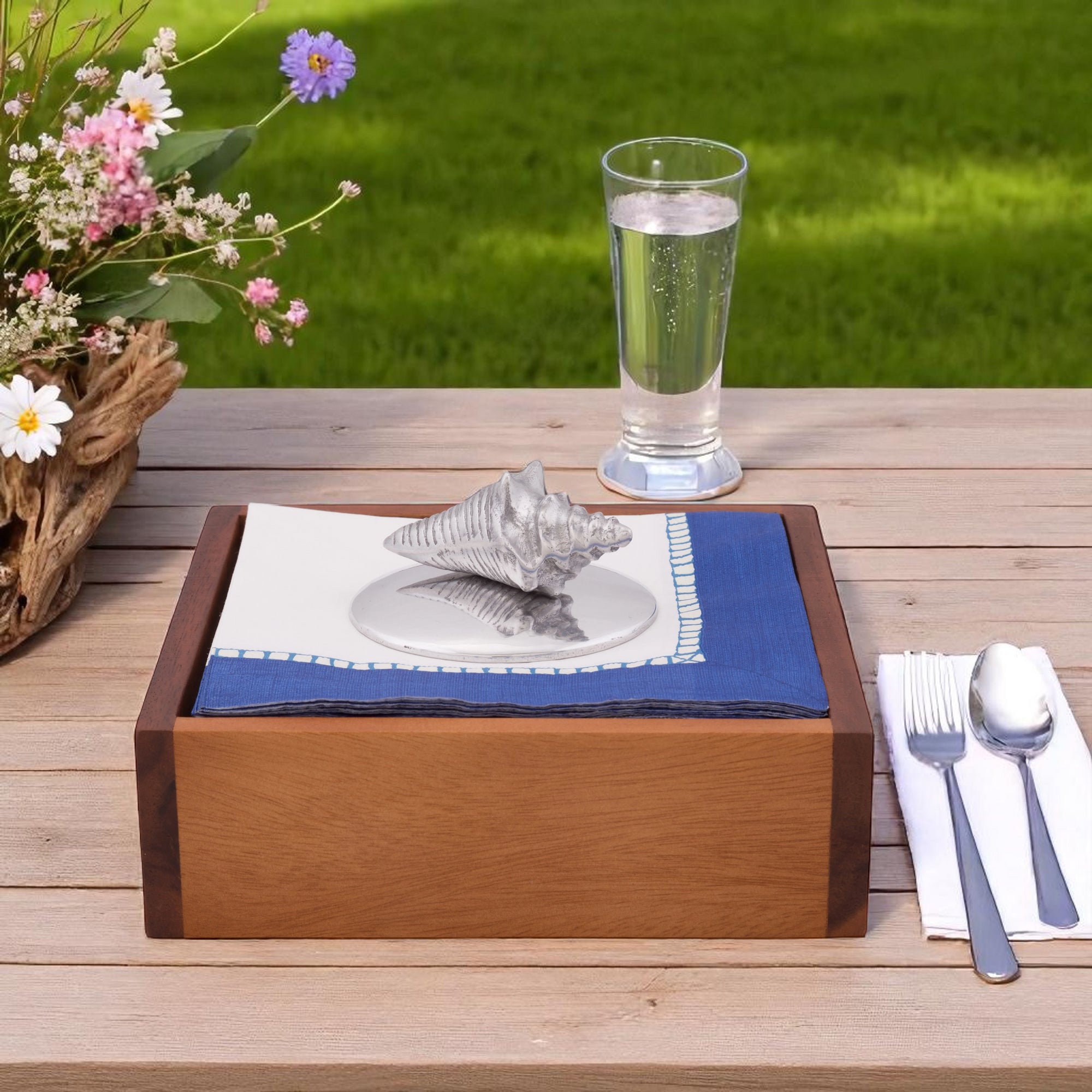A coastal decor napkin weight featuring a silver shell design atop blue and white napkins, viewed from the front angle.