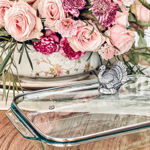 A casserole server featuring a turkey-shaped handle viewed from the side, set against a bouquet of pink roses.