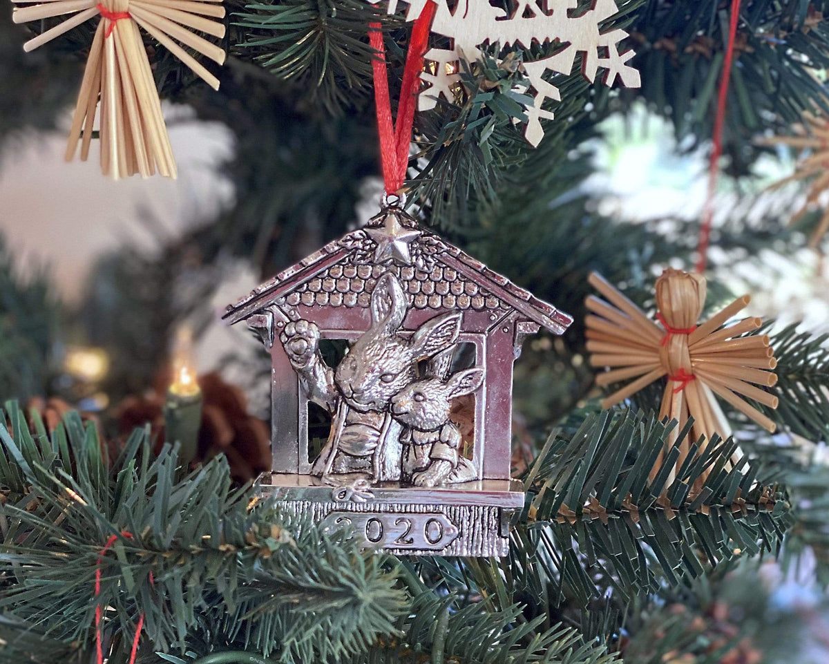 A charming Christmas ornament featuring rabbits inside a house is angled amidst tree branches, surrounded by festive decor.