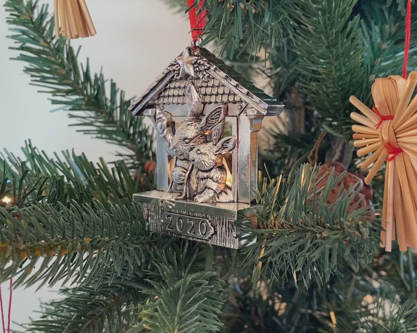 A charming Christmas ornament featuring rabbits inside a house is angled amidst tree branches, surrounded by festive decor.